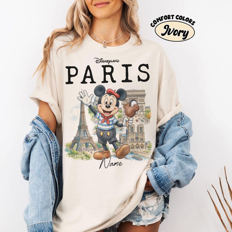 K&ouml;nnte beinhalten: Elfenbeinfarbenes T-Shirt mit dem schwarzen Aufdruck "Disneyland PARIS". Es zeigt eine Cartoon-Mickey-Mouse in Matrosenkleidung, die ein Schokoladeneis h&auml;lt und vor dem Eiffelturm und anderen Pariser Wahrzeichen winkt. Das Shirt ist mit "Comfort Colors" gekennzeichnet.
