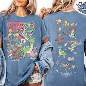 May include: Blue jean-colored t-shirt featuring a colorful graphic of Toy Story characters. The front of the shirt displays Jessie, Woody, Buzz, Rex, Hamm, and Slinky Dog. The back of the shirt shows additional characters and the Toy Story logo.