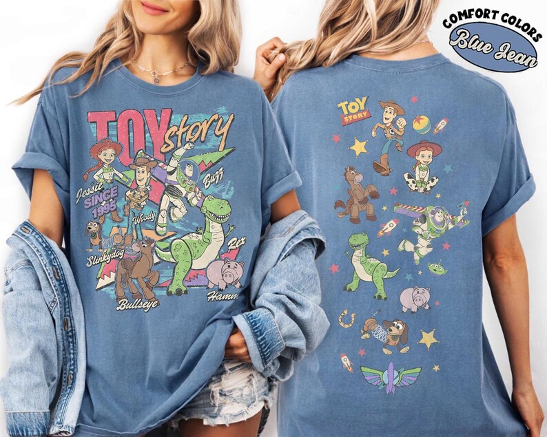 May include: Blue jean-colored t-shirt featuring a colorful graphic of Toy Story characters. The front of the shirt displays Jessie, Woody, Buzz, Rex, Hamm, and Slinky Dog. The back of the shirt shows additional characters and the Toy Story logo.