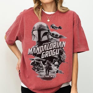 Op de afbeelding: Rood getint T-shirt met een grafisch ontwerp uit de Star Wars-serie, "The Mandalorian and Grogu". Het ontwerp bevat afbeeldingen van The Mandalorian, Grogu en de tekst "This is the Way".