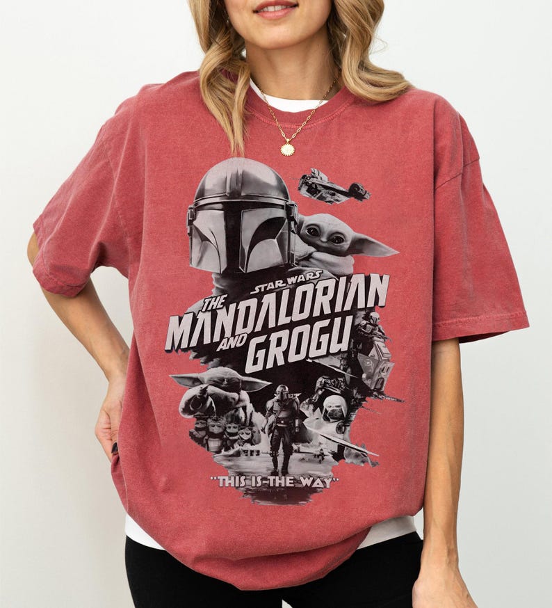 Op de afbeelding: Rood getint T-shirt met een grafisch ontwerp uit de Star Wars-serie, "The Mandalorian and Grogu". Het ontwerp bevat afbeeldingen van The Mandalorian, Grogu en de tekst "This is the Way".