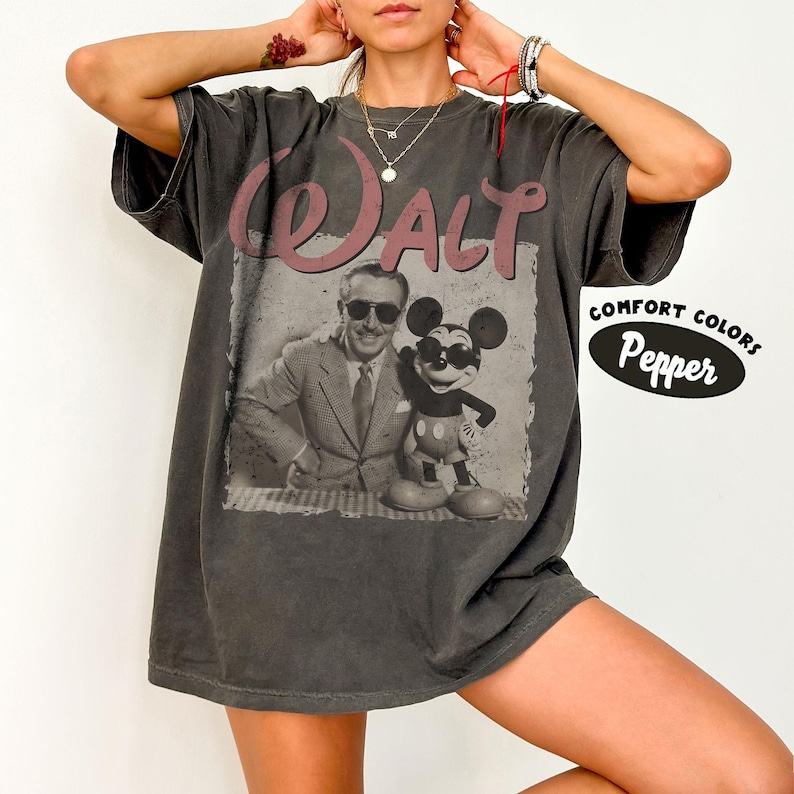 May include: A gray t-shirt featuring a vintage-style graphic of Walt Disney and Mickey Mouse. The word "Walt" is printed in a pinkish-red color above the image. The shirt also has the words "Comfort Colors Pepper" printed on the right side.