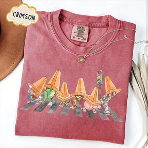 May include: Crimson-colored t-shirt with a graphic of cartoon characters crossing a street, wearing orange traffic cones. The shirt has a small Comfort Colors tag and a silver necklace. The word "CRIMSON" is in a cloud-shaped text box.