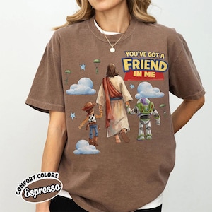 May include: Espresso-coloured t-shirt with a graphic featuring Jesus holding hands with Woody and Buzz Lightyear. The text "YOU'VE GOT A FRIEND IN ME" is displayed above the characters. The shirt has a crew neck and short sleeves.
