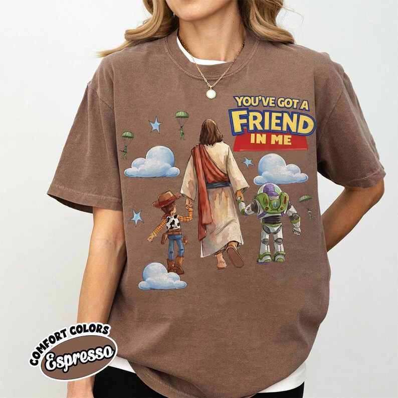 May include: Espresso-coloured t-shirt with a graphic featuring Jesus holding hands with Woody and Buzz Lightyear. The text "YOU'VE GOT A FRIEND IN ME" is displayed above the characters. The shirt has a crew neck and short sleeves.