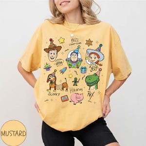 Pu&ograve; includere: T-shirt color senape con illustrazioni di personaggi dei cartoni animati di Toy Story, tra cui Woody, Buzz Lightyear, Jessie, Slinky, Hamm e Rex. Il design include anche altri elementi. La maglietta ha una vestibilit&agrave; ampia.