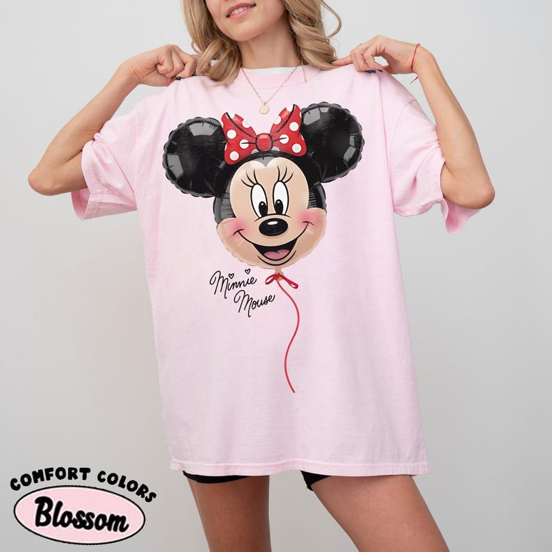 K&ouml;nnte beinhalten: Hellrosa T-Shirt mit einem Minnie Mouse Ballon-Design. Der Ballon hat schwarze Ohren, eine rote Schleife mit wei&szlig;en Punkten und ein l&auml;chelndes Gesicht. Die Worte "Minnie Mouse" sind in Kursivschrift unter dem Ballon geschrieben.