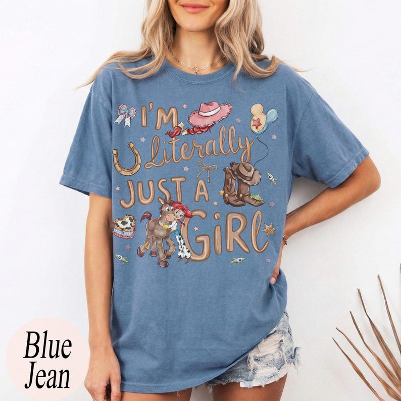 K&ouml;nnte beinhalten: Blaues Jeans-T-Shirt mit dem Text "I'm Literally Just a Girl" und Cartoon-Grafiken eines Cowboyhuts, Stiefeln, eines Pferdes und anderer Elemente. Das Shirt hat eine lockere Passform.