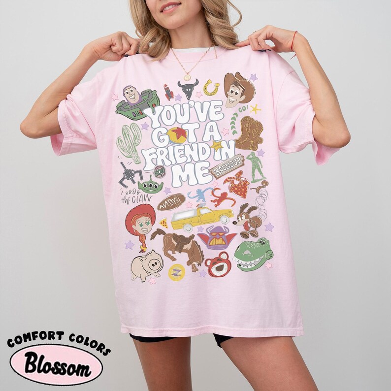Op de afbeelding: Een lichtroze T-shirt met korte mouwen en een kleurrijke grafische print met de tekst "YOU'VE GOT A FRIEND IN ME", omringd door diverse illustraties van stripfiguren, waaronder een cowboy, een space ranger en een dinosaurus.