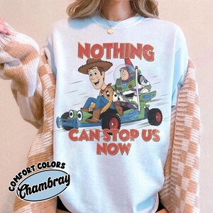 May include: A light blue crewneck sweatshirt featuring a graphic of Woody and Buzz Lightyear riding a green go-kart. The text above and below the characters reads NOTHING CAN STOP US NOW in a distressed red font. A Comfort Colors Chambray label is visible.