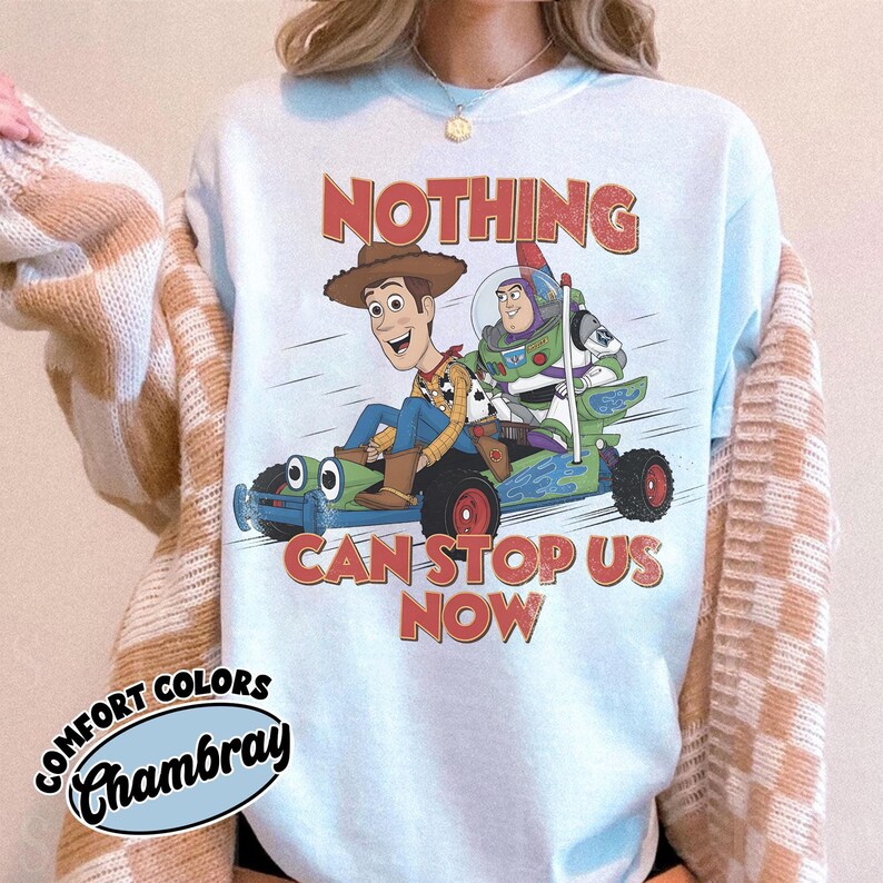 May include: A light blue crewneck sweatshirt featuring a graphic of Woody and Buzz Lightyear riding a green go-kart. The text above and below the characters reads NOTHING CAN STOP US NOW in a distressed red font. A Comfort Colors Chambray label is visible.