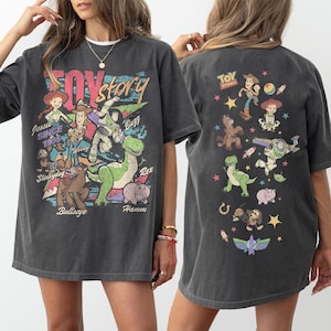 May include: Dark gray t-shirt featuring a colorful graphic of Toy Story characters, including Woody, Buzz Lightyear, and Rex. The front of the shirt displays the words "Toy Story" and "Since 1995". The back of the shirt also has a Toy Story graphic.