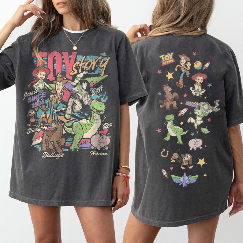 May include: Dark gray t-shirt featuring a colorful graphic of Toy Story characters, including Woody, Buzz Lightyear, and Rex. The front of the shirt displays the words "Toy Story" and "Since 1995". The back of the shirt also has a Toy Story graphic.