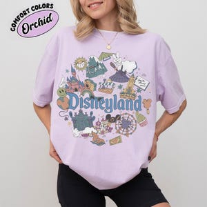 May include: A light purple t-shirt with a Disneyland graphic. The graphic features various Disneyland icons, including a castle, a Ferris wheel, and a smiling sun. The text "Disneyland" is in blue. The shirt has the text "Comfort Colors Orchid" in a circle.