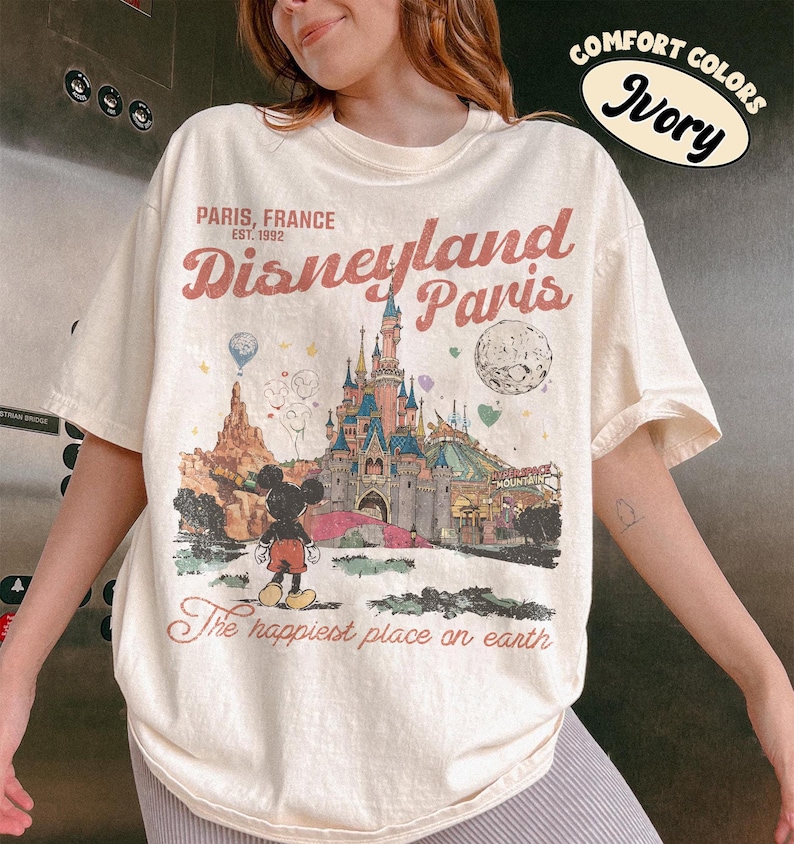 May include: Ivory-colored t-shirt featuring a Disneyland Paris graphic. The design includes a castle, Mickey Mouse, and the text "PARIS, FRANCE Disneyland Paris The happiest place on earth". The shirt is a comfort colors style.