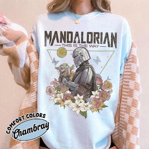 May include: Light blue sweatshirt featuring a graphic of The Mandalorian and Grogu surrounded by flowers and butterflies. The text "MANDALORIAN - THIS IS THE WAY" is printed across the top. The sweatshirt is labeled "Comfort Colors Chambray".