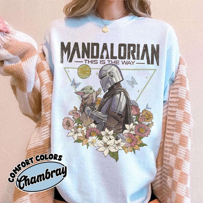 May include: Light blue sweatshirt featuring a graphic of The Mandalorian and Grogu surrounded by flowers and butterflies. The text "MANDALORIAN - THIS IS THE WAY" is printed across the top. The sweatshirt is labeled "Comfort Colors Chambray".