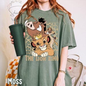 May include: A moss green t-shirt featuring a graphic of characters from The Lion King. The design includes Simba wearing a crown, Pumbaa, and Timon. The words "THE LION KING" are printed below the characters. A dark green tumbler is held in the left hand.