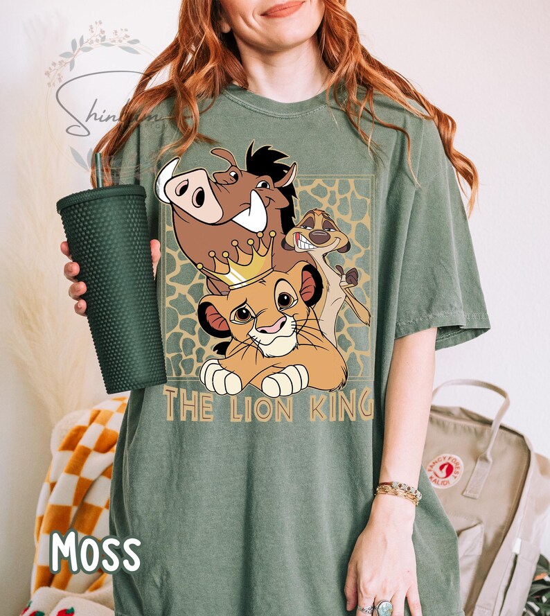 May include: A moss green t-shirt featuring a graphic of characters from The Lion King. The design includes Simba wearing a crown, Pumbaa, and Timon. The words "THE LION KING" are printed below the characters. A dark green tumbler is held in the left hand.