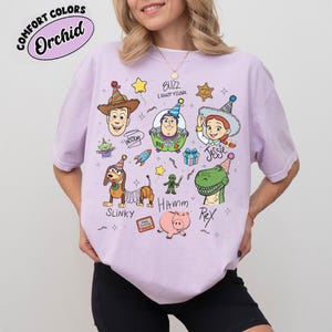 Pu&ograve; includere: Una maglietta lilla con personaggi dei cartoni animati del film Toy Story. La maglietta presenta Woody, Buzz Lightyear, Jessie, Slinky, Hamm e Rex. Il testo "COMFORT COLORS" e "Orchid" &egrave; in un cerchio in alto a sinistra.