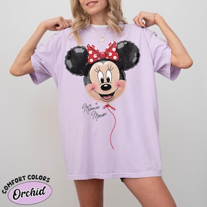 K&ouml;nnte beinhalten: Ein helllila T-Shirt mit einem Minnie Mouse Ballon-Design. Der Ballon hat schwarze Ohren, eine rote Schleife mit wei&szlig;en Punkten und ein l&auml;chelndes Gesicht. Die Worte "Minnie Mouse" stehen darunter.