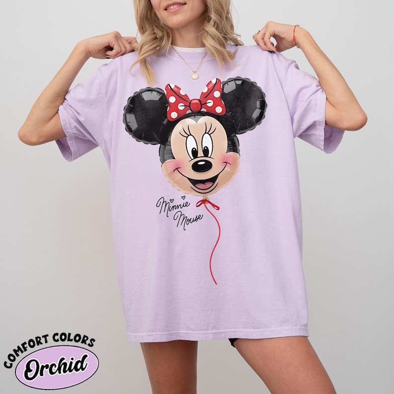K&ouml;nnte beinhalten: Ein helllila T-Shirt mit einem Minnie Mouse Ballon-Design. Der Ballon hat schwarze Ohren, eine rote Schleife mit wei&szlig;en Punkten und ein l&auml;chelndes Gesicht. Die Worte "Minnie Mouse" stehen darunter.