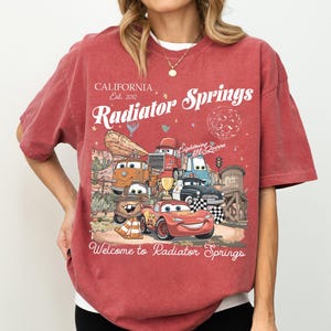 以下が含まれることがあります： 映画「カーズ」のキャラクターのグラフィックが特徴の赤いTシャツ。テキストには「California Est. 2012 Radiator Springs」と「Welcome to Radiator Springs」が含まれています。ヴィンテージ感のあるデザインです。
