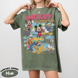 Peut inclure: T-shirt vert mousse avec un motif de Mickey Mouse et ses amis. Le motif comprend Mickey, Minnie, Donald Duck, Dingo, Pluto et Daisy Duck. Le texte indique "Mickey & Friends" et "Since 1928". Le t-shirt est &eacute;tiquet&eacute; "Comfort Colors".