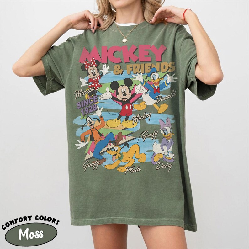 Peut inclure: T-shirt vert mousse avec un motif de Mickey Mouse et ses amis. Le motif comprend Mickey, Minnie, Donald Duck, Dingo, Pluto et Daisy Duck. Le texte indique "Mickey & Friends" et "Since 1928". Le t-shirt est &eacute;tiquet&eacute; "Comfort Colors".
