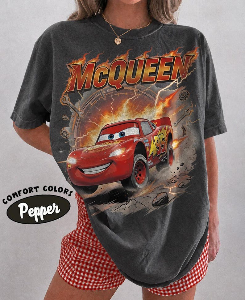 Może przedstawiać: Ciemnoszara koszulka z grafiką Zygzaka McQueena z filmu Auta. Projekt zawiera napis "McQueen" w pomarańczowych i czerwonych płomieniach oraz numer "95" na samochodzie. Koszulka ma napisy "Comfort Colors" i "Pepper" po lewej stronie u dołu.