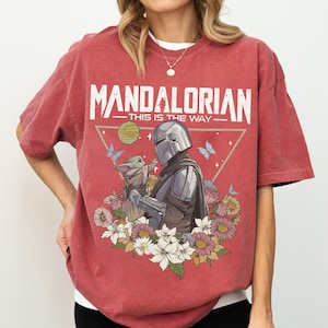 May include: Red t-shirt featuring the text "MANDALORIAN" above an illustration of the Mandalorian and Grogu surrounded by flowers and butterflies. The phrase "THIS IS THE WAY" is below the text.