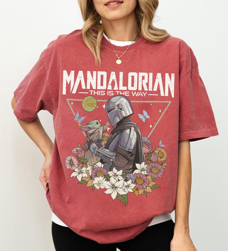 May include: Red t-shirt featuring the text "MANDALORIAN" above an illustration of the Mandalorian and Grogu surrounded by flowers and butterflies. The phrase "THIS IS THE WAY" is below the text.