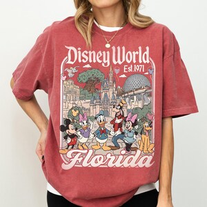 May include: Red t-shirt featuring a Disney World Florida graphic with Mickey Mouse, Minnie Mouse, Donald Duck, Daisy Duck, Goofy, and Pluto. The graphic includes the text "Disney World Est. 1971" and "Florida".
