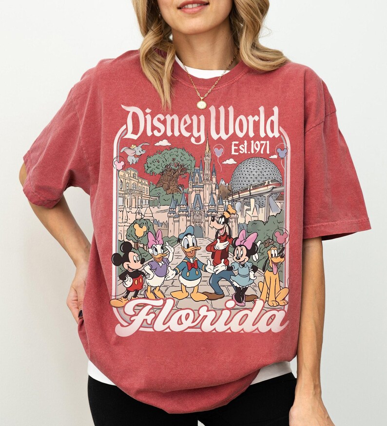 May include: Red t-shirt featuring a Disney World Florida graphic with Mickey Mouse, Minnie Mouse, Donald Duck, Daisy Duck, Goofy, and Pluto. The graphic includes the text "Disney World Est. 1971" and "Florida".