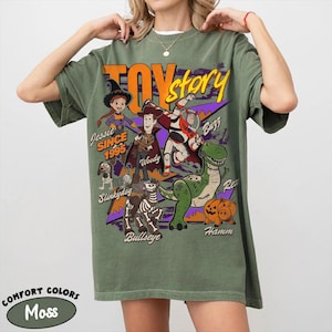 May include: A moss green t-shirt featuring a graphic of characters from the movie "Toy Story." The design includes Woody, Buzz Lightyear, Jessie, Rex, Hamm, Slinkydog, and Bullseye. The text "Toy Story" and "Since 1995" are also visible.