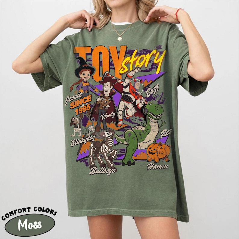 May include: A moss green t-shirt featuring a graphic of characters from the movie "Toy Story." The design includes Woody, Buzz Lightyear, Jessie, Rex, Hamm, Slinkydog, and Bullseye. The text "Toy Story" and "Since 1995" are also visible.