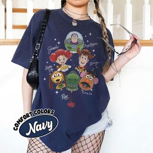 Puede incluir: Camiseta azul marino con un gr&aacute;fico de personajes de Toy Story, incluyendo a Buzz Lightyear, Woody y Jessie. La camiseta tiene el texto "Comfort Colors Navy" en un &oacute;valo blanco. El dise&ntilde;o incluye estrellas y nombres de personajes.