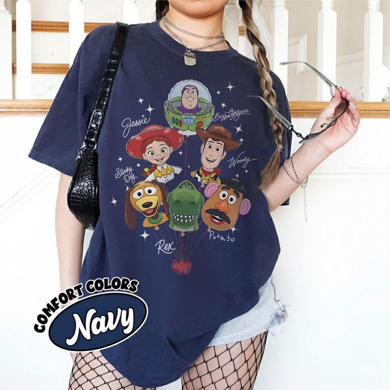 Puede incluir: Camiseta azul marino con un gr&aacute;fico de personajes de Toy Story, incluyendo a Buzz Lightyear, Woody y Jessie. La camiseta tiene el texto "Comfort Colors Navy" en un &oacute;valo blanco. El dise&ntilde;o incluye estrellas y nombres de personajes.