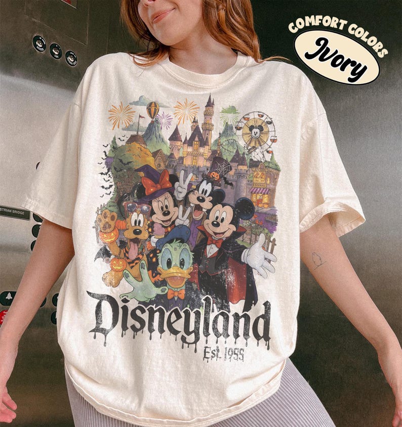 May include: An ivory short-sleeve t-shirt featuring a graphic print of Disney characters in Halloween costumes, including Mickey, Minnie, Donald, and Pluto in front of a castle. The text reads Disneyland Est. 1955 and Comfort Colors Ivory.