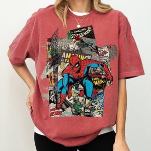 Pode incluir: Camiseta vermelha com um design de colagem de hist&oacute;rias em quadrinhos com o Homem-Aranha. O design incorpora capas de hist&oacute;rias em quadrinhos com o texto "AMAZING SPIDER-MAN" e "ZING MAN". A camiseta &eacute; de cor vermelha lisa.