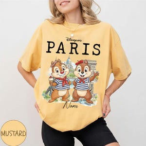 K&ouml;nnte beinhalten: Senffarbenes T-Shirt mit dem Text "Disneyland PARIS" &uuml;ber einer Grafik von zwei Comic-Chipmunks vor Pariser Wahrzeichen. Die Chipmunks tragen Matrosenhemden und Baretts. Das Wort "Name" steht darunter.