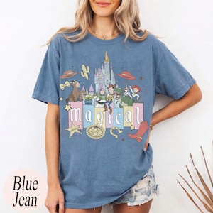 Puede incluir: Camiseta azul vaquero con un gr&aacute;fico colorido con la palabra "m&aacute;gico" y personajes de dibujos animados. El dise&ntilde;o incluye un castillo, sombrero de vaquero, cactus y una placa de sheriff.