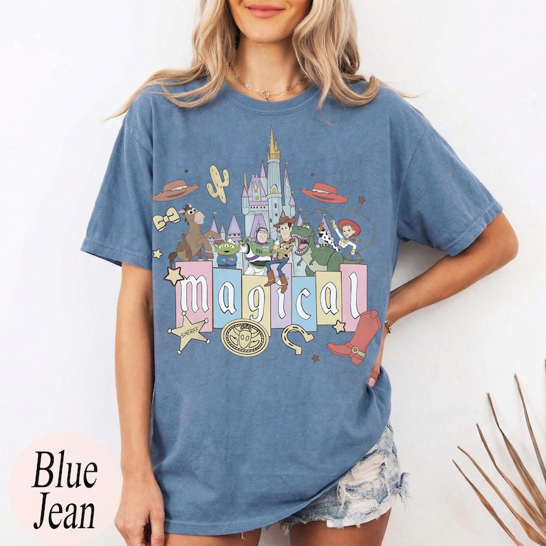 Puede incluir: Camiseta azul vaquero con un gr&aacute;fico colorido con la palabra "m&aacute;gico" y personajes de dibujos animados. El dise&ntilde;o incluye un castillo, sombrero de vaquero, cactus y una placa de sheriff.