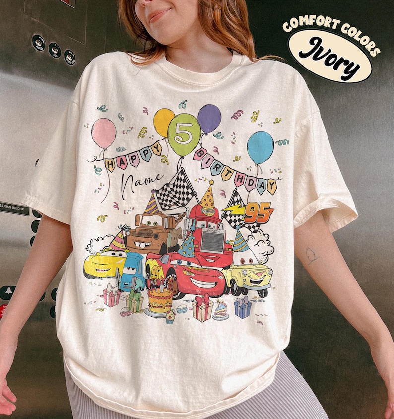 May include: Ivory-colored t-shirt with a colorful graphic featuring cartoon cars, balloons, and the text "HAPPY BIRTHDAY" and "95". The design includes a birthday cake and gift boxes, perfect for a fifth birthday celebration.