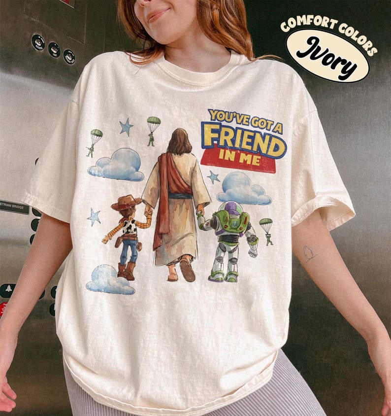May include: Ivory-coloured t-shirt with the text "YOU'VE GOT A FRIEND IN ME" and a graphic of Jesus, Woody, and Buzz Lightyear. The shirt features a crew neck and short sleeves. The words "Comfort Colors" and "Ivory" are printed on the top right.