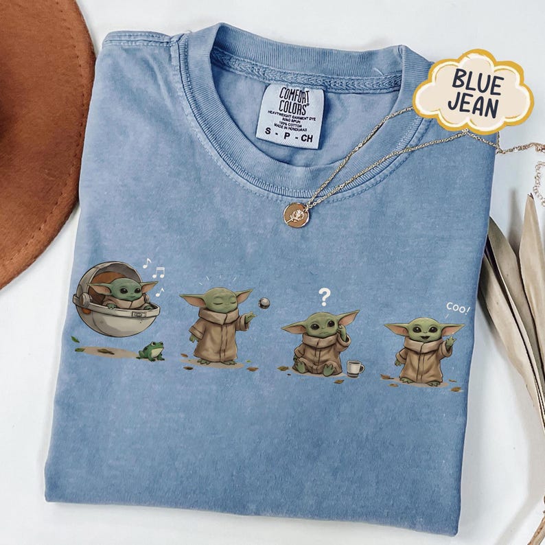 Puede incluir: Camiseta azul vaquero claro con un gr&aacute;fico de un personaje en varias poses. El personaje est&aacute; en una c&aacute;psula, jugando con una pelota, confundido y diciendo "coo!". Un collar dorado est&aacute; sobre la camiseta.