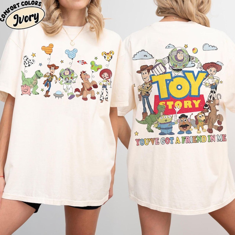 May include: An ivory t-shirt featuring Toy Story characters. The front displays Woody, Buzz Lightyear, Rex, Bullseye, Jessie, and Hamm with Mickey-shaped balloons. The back shows the Toy Story logo and the text You've Got A Friend In Me.