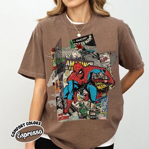 Pode incluir: Camiseta marrom com um design de colagem de quadrinhos do Homem-Aranha. O design inclui as palavras "The Amazing Spider-Man" e "Zing Man". A camiseta &eacute; da marca "Comfort Colors".