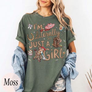 K&ouml;nnte beinhalten: Moosgr&uuml;nes T-Shirt mit dem Text "I'm Literally Just a Girl" und einer Grafik eines Hufeisens, Cowboyhuts, Stiefel und eines Cartoon-Pferdes und Cowgirls. Das Shirt hat kurze &Auml;rmel und eine lockere Passform.