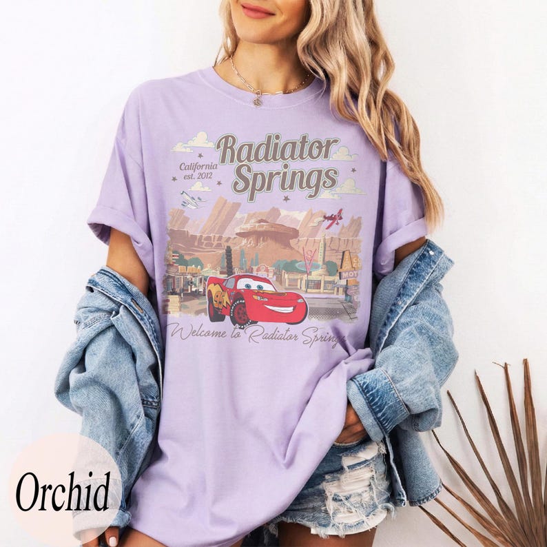 May include: A lilac t-shirt with a graphic of Radiator Springs from the film Cars. The design includes the text "Radiator Springs" and "Welcome to Radiator Spring" with an image of Lightning McQueen. The shirt has a loose fit.